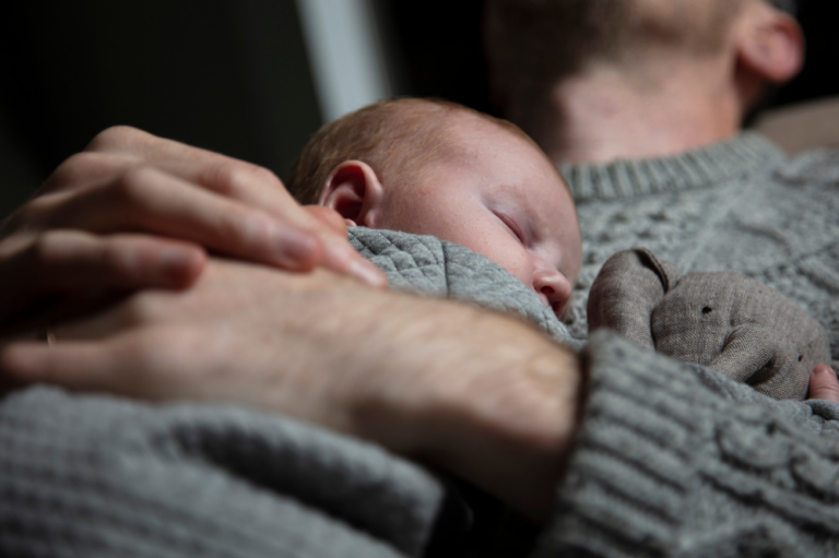 Vater in grauem Strickpullover hält sein schlafendes Neugeborenes an der Brust, daneben ein kleines Kuscheltier.